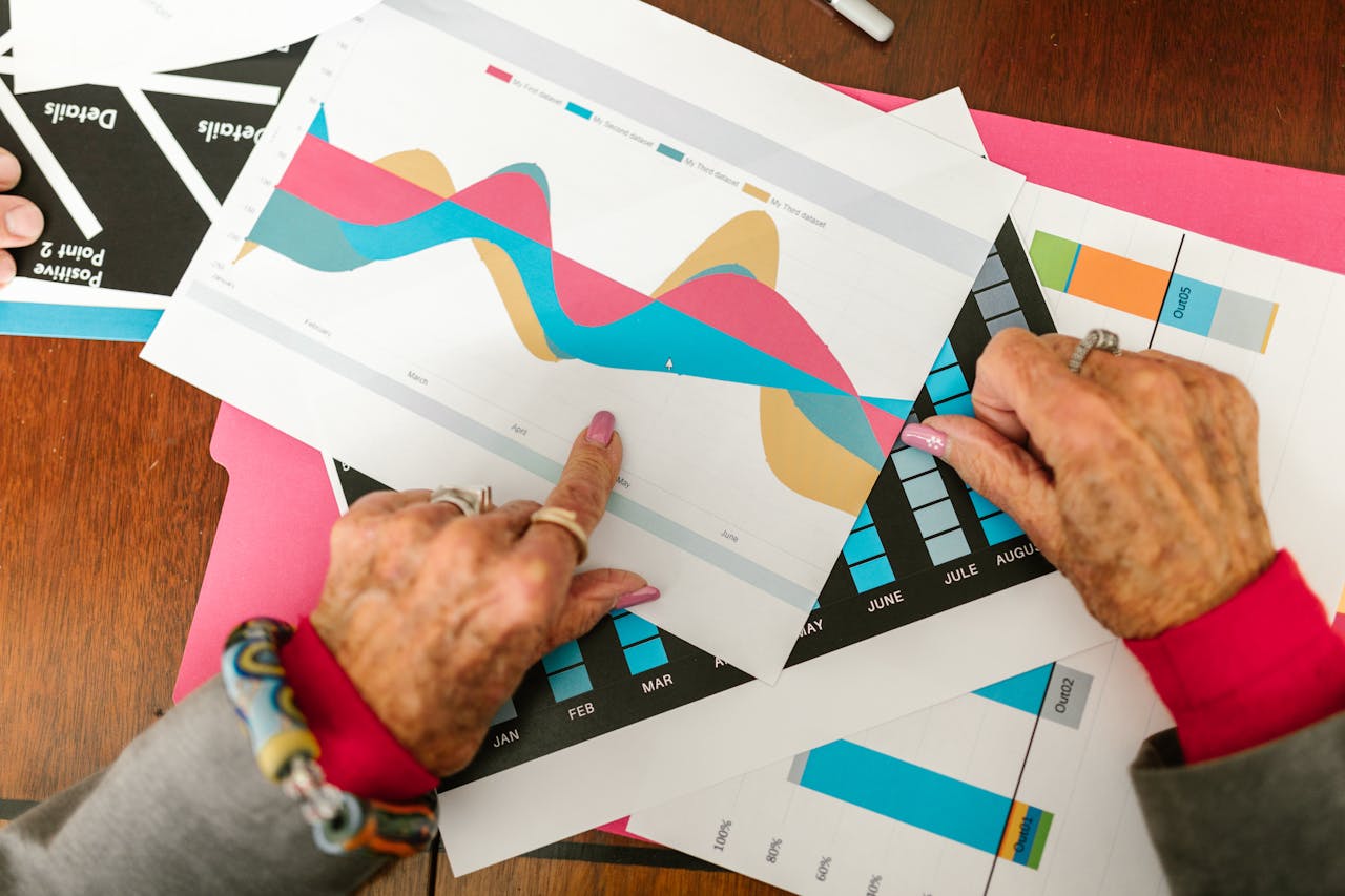 Senior adult pointing at colorful business graphs with data analysis documents on a wooden table.