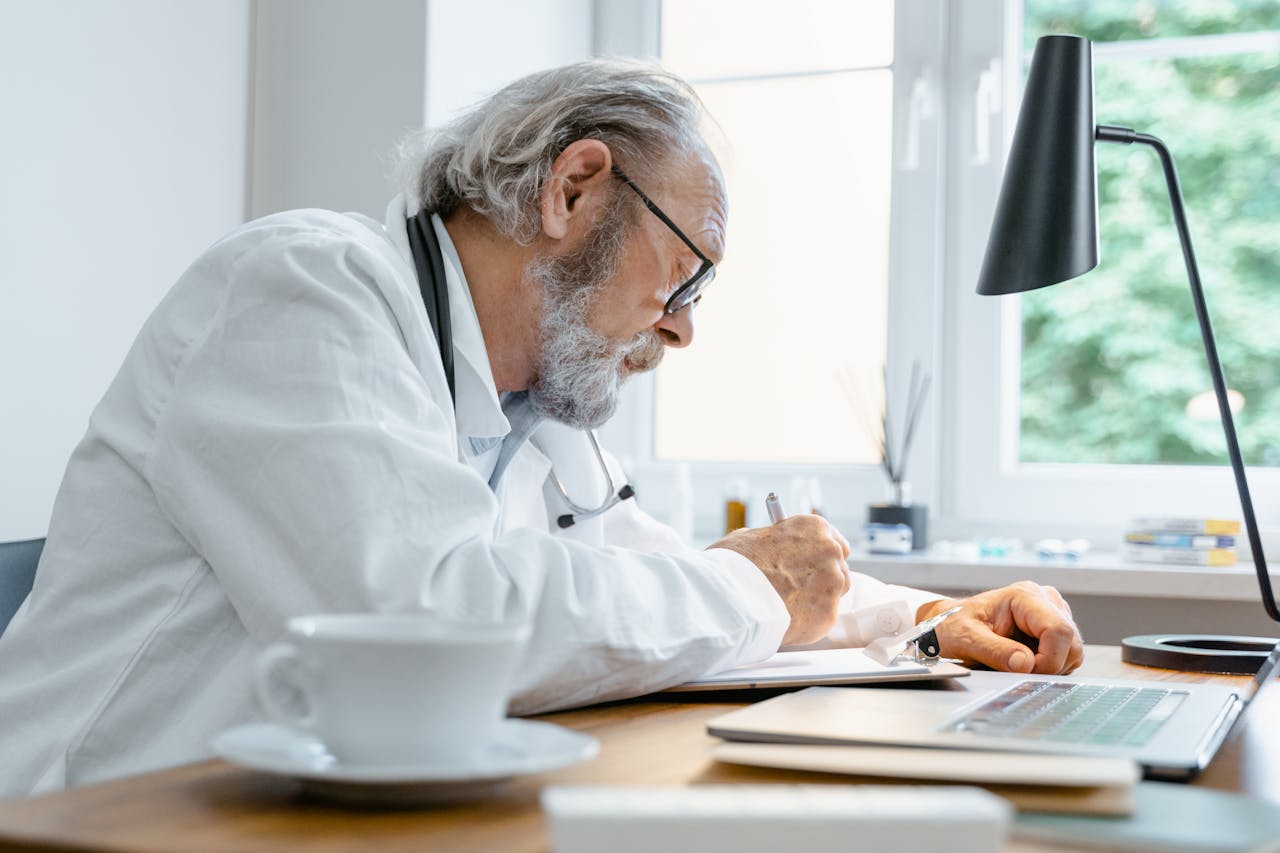 Elderly doctor writing notes at his desk with a focus on work in a well-lit office.