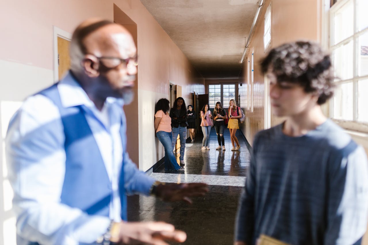A blurred teacher talks to a student in a school hallway, with peers in the background.